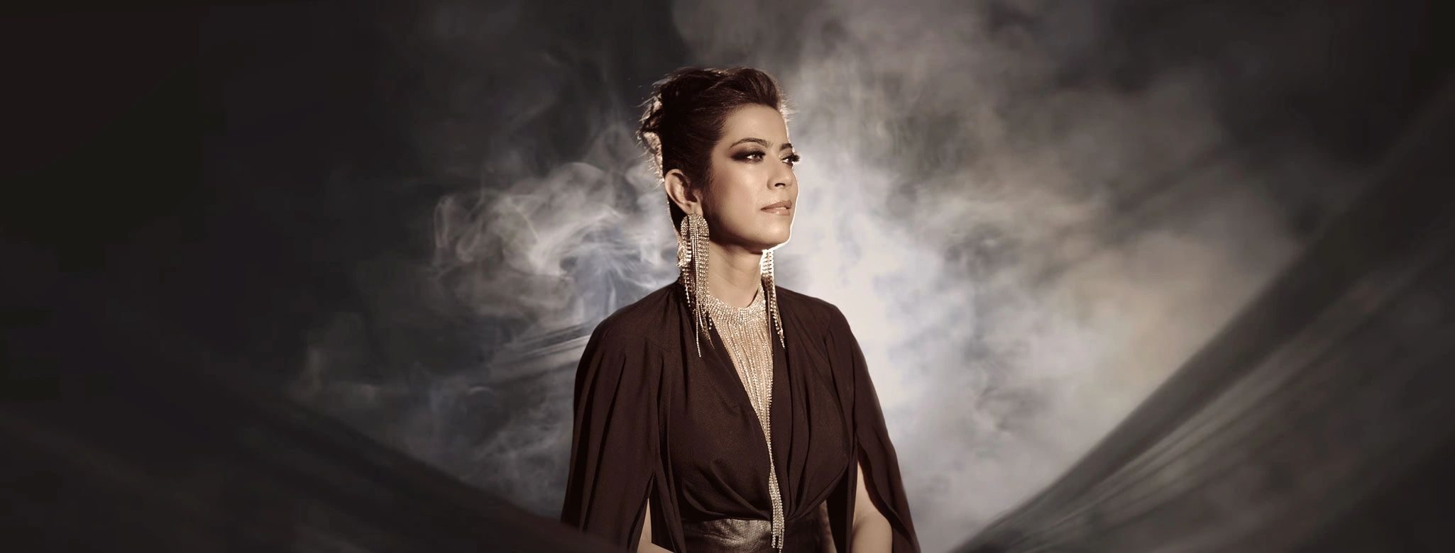 Devika in a dark cape with long gold earrings, looking to the side through drifting smoke.