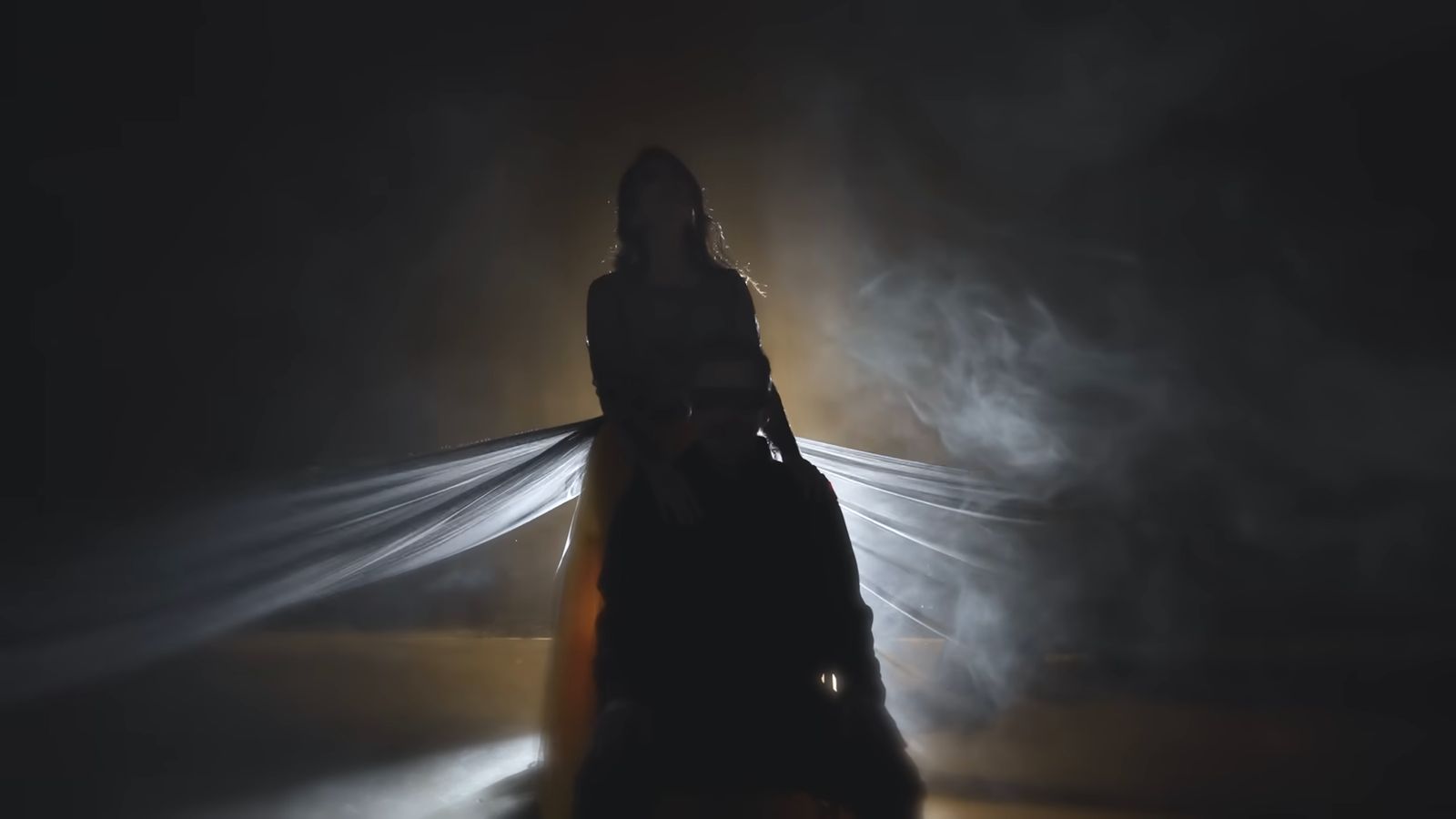 Devika in silhouette, arms outstretched, veils lit from behind in smoke.