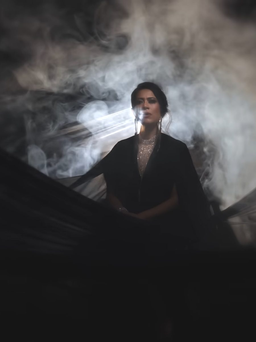 Devika standing in drifting smoke with a sheer black veil lifted around her.